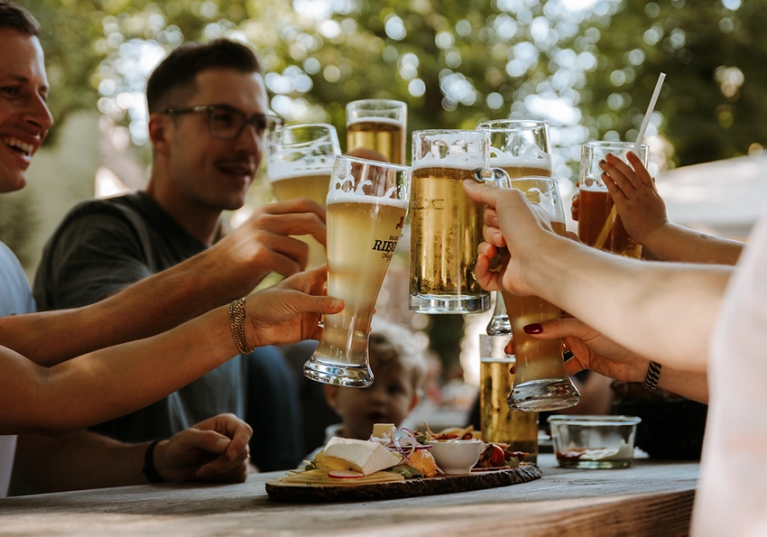 Eine Gruppe Menschen die mit Biergläsern anstoßt. Auf dem Tisch steht ein Brotzeitbrett.